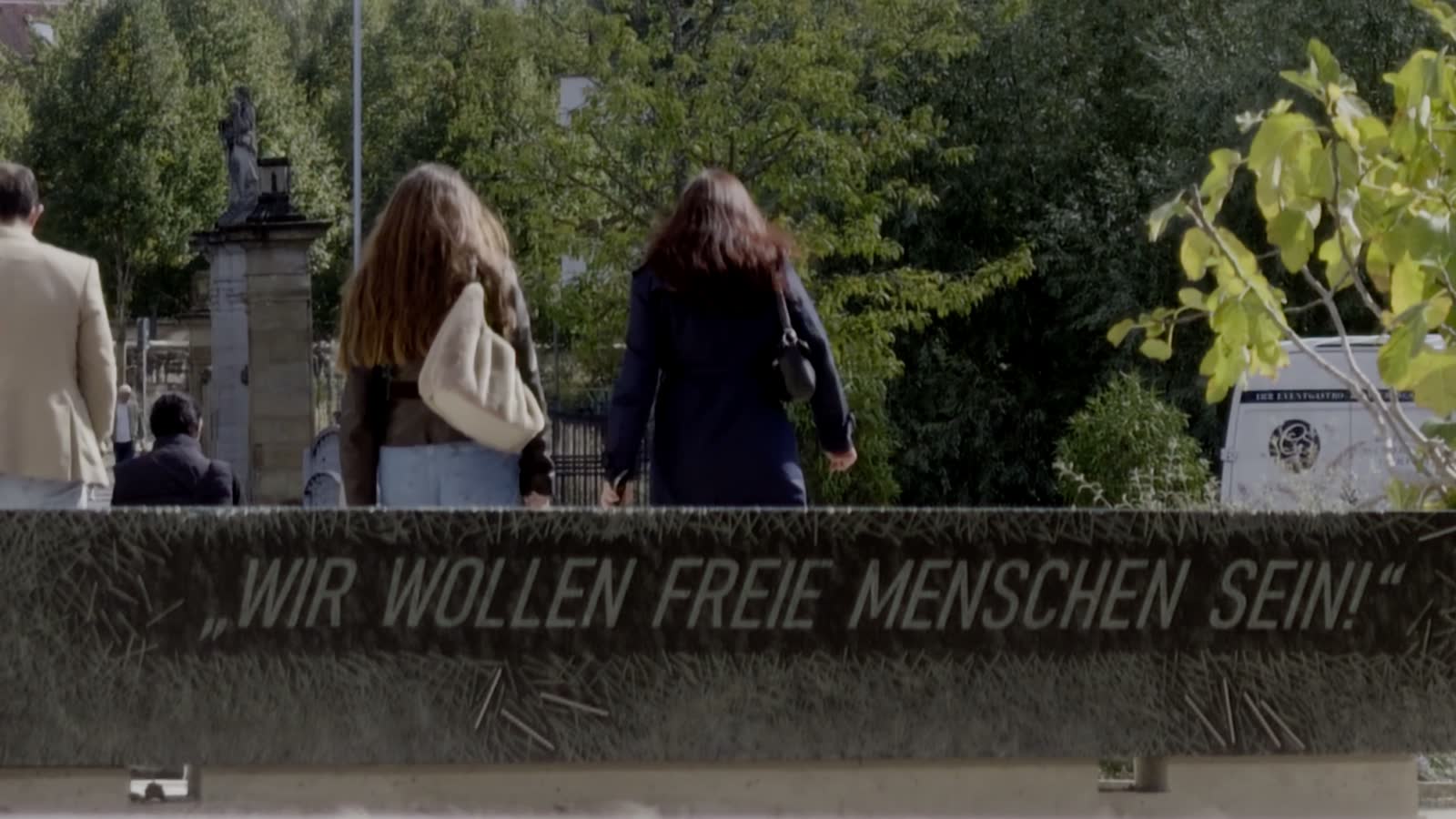 Nahaufnahme der Inschrift „Wir wollen freie Menschen sein!“ auf der Längsseite des Gedenksteins; dahinter gehen zwei junge Frauen vorbei.